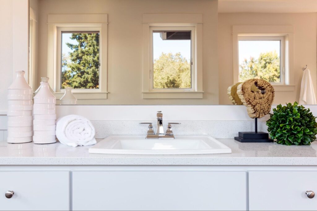 A beautiful bathroom with neutral colors featuring a sink, mirrors reflecting outdoor trees, and elegant decor accents designed by professional remodelers.