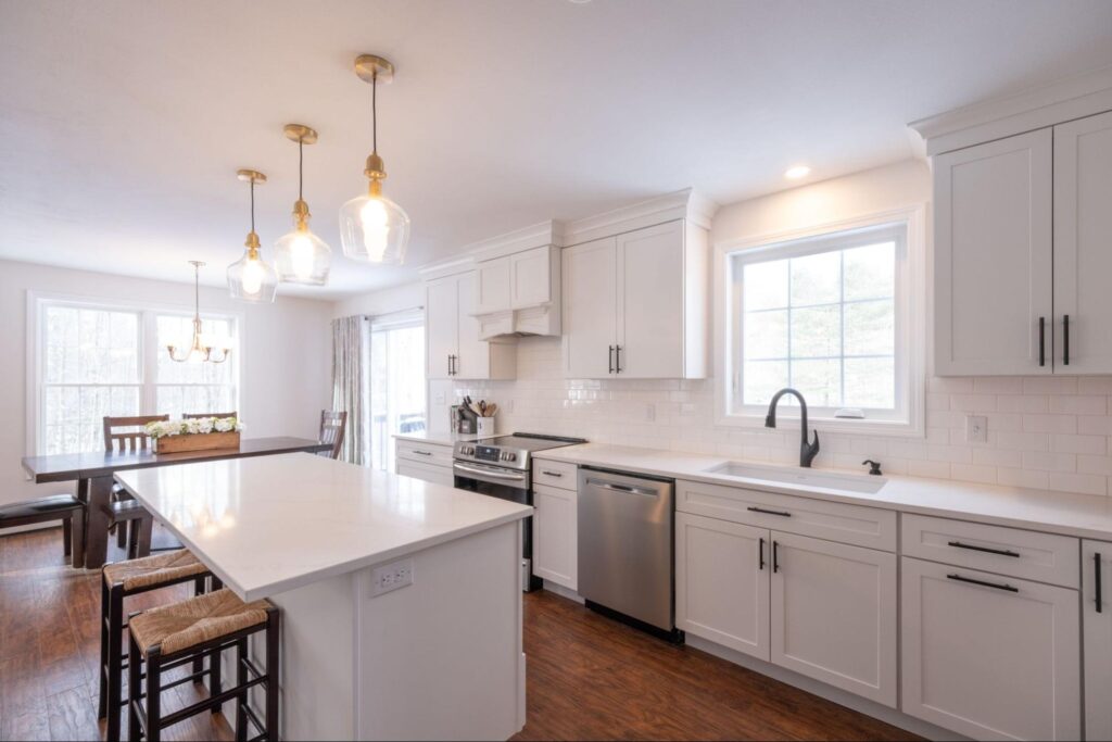 A newly remodeled modern white kitchen interior featuring an oven, sink, faucet, wooden cabinets, and table, with hanging lamps above the kitchen island.