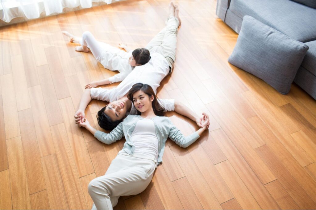 Man, woman, and child lying down on a wooden floor next to a pillow and a couch.