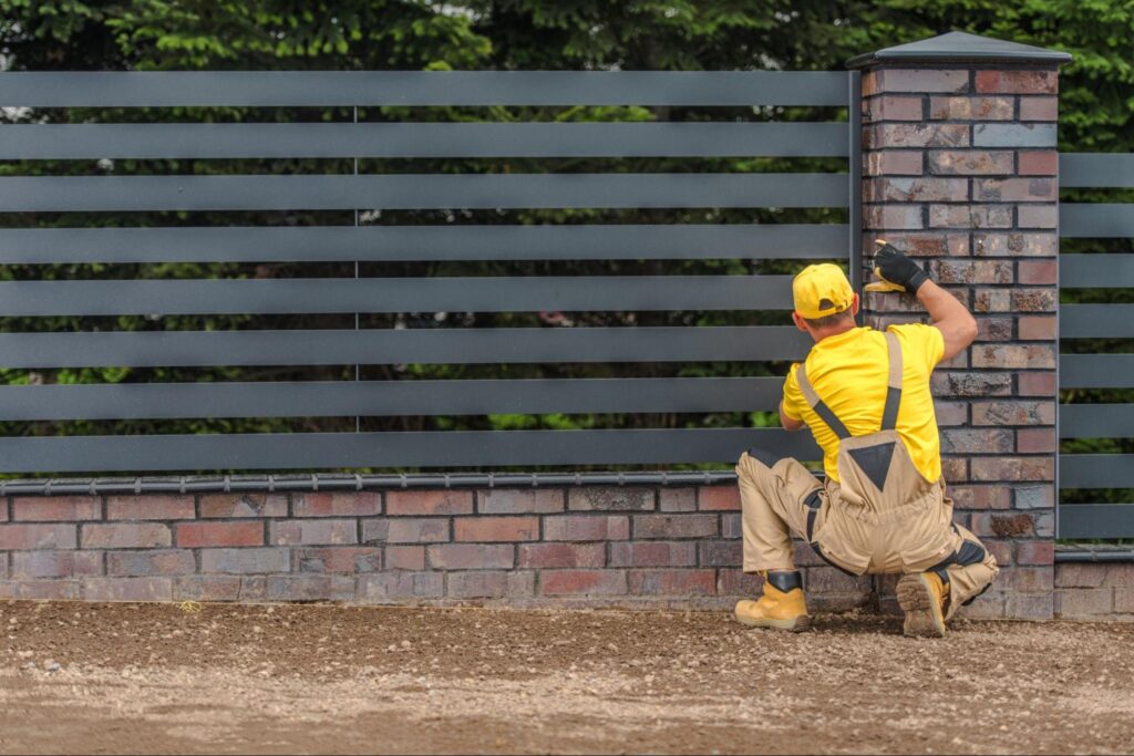 A professional fence installer in a yellow and beige outfit completes the installation of metal panels between concrete brick posts.