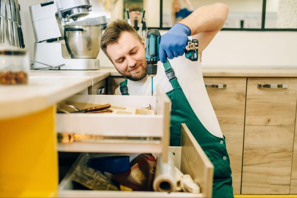 A handyman in a green uniform drives screws into an upper kitchen cabinet while the lower cabinet door is open.