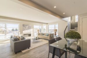 A spacious living room with an open floor plan featuring large windows that fill the space with natural light, modern furniture including a gray and beige sofa set, a glass dining table adorned with a green plant centerpiece, and a cozy area rug.